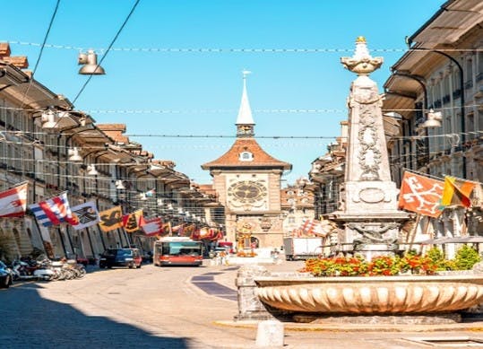 A street scene with a historic clock tower, lined with flags and buildings, featuring a decorative fountain in the foreground.
