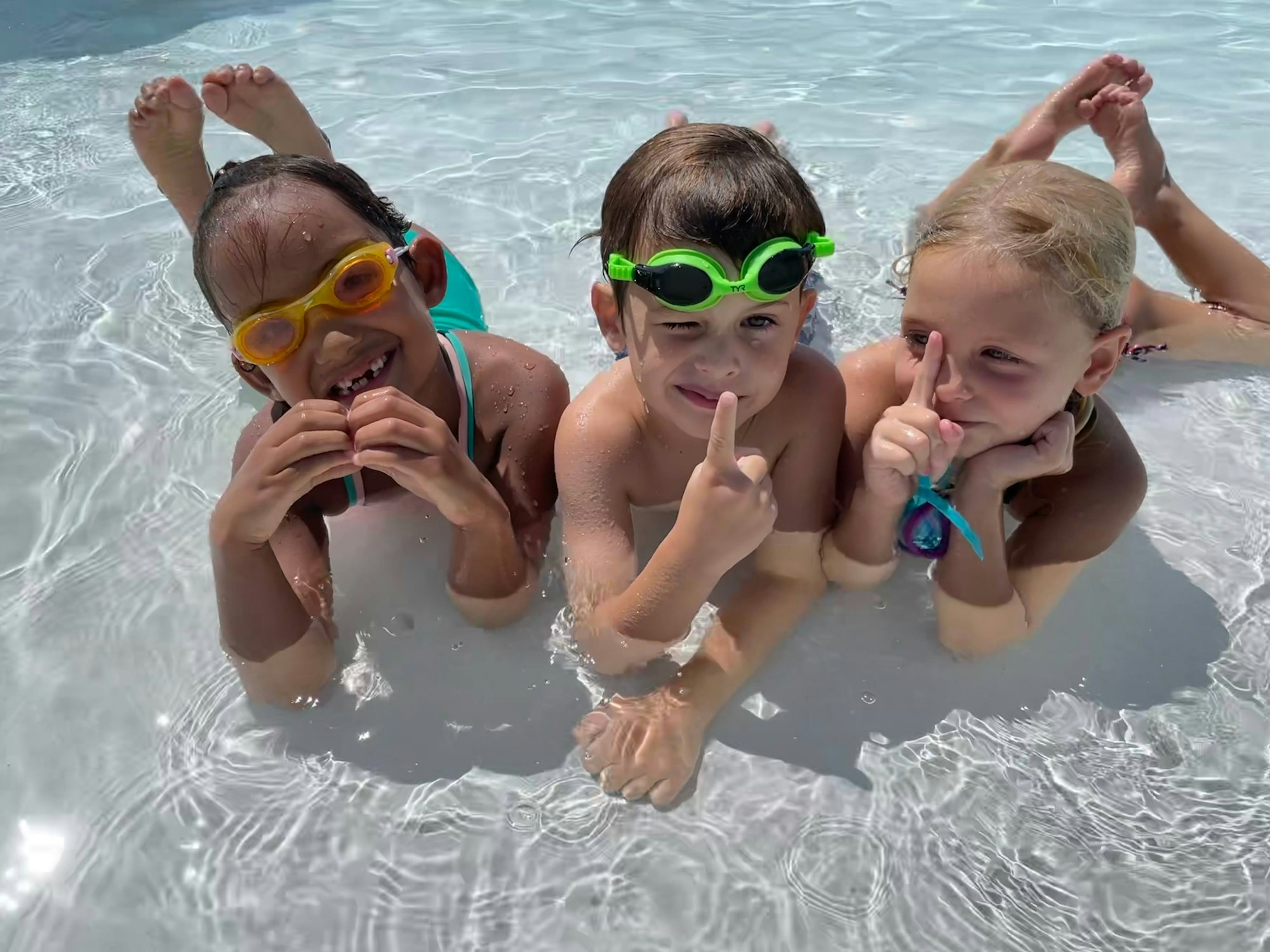 Three children with swim goggles, smiling in a pool, making playful gestures with their hands.