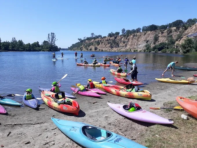 The image shows a group of people kayaking and paddleboarding by a river, with colorful kayaks lined up on the shore.