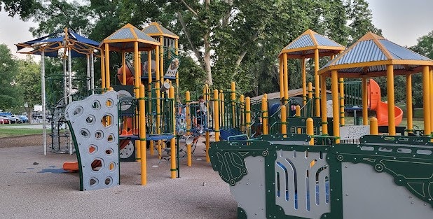 The image shows a colorful playground with various play structures, slides, and climbing features in a park setting.