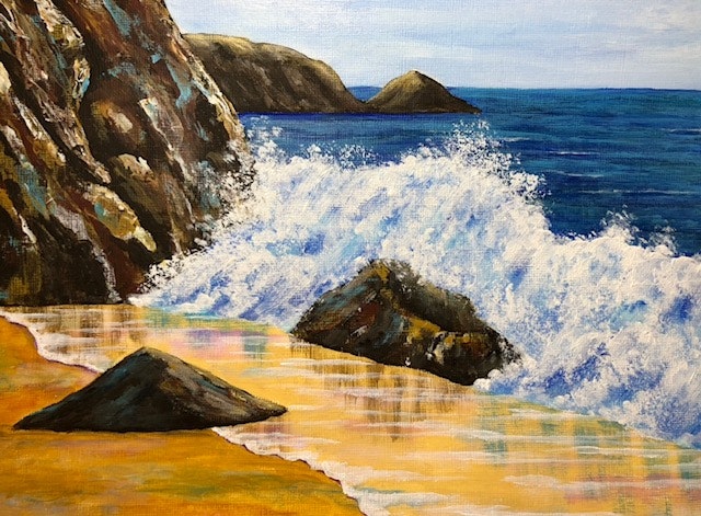 Waves crashing against rocks on a sandy beach with cliffs and distant hills under a blue sky.