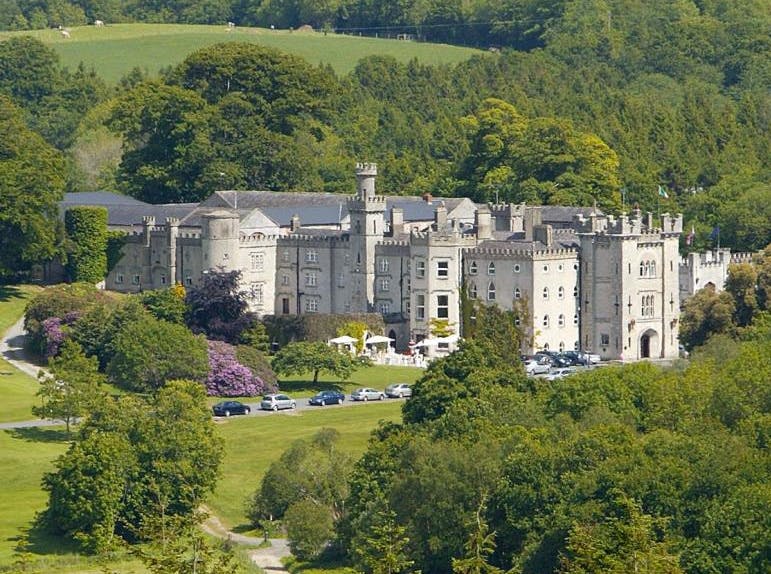 A large castle surrounded by trees and green fields, with a few cars parked in front.