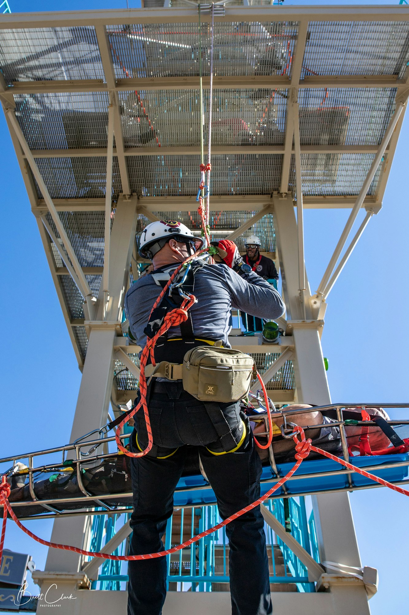 A person in climbing gear is performing a rescue operation with ropes, near a stretcher on a metal structure.