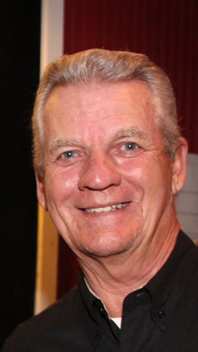 A smiling older man in a black shirt, posing against a blurred background.