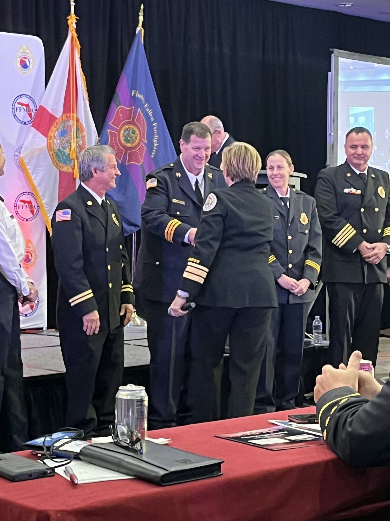 A group of people in formal uniforms stands on a stage with flags and conference materials displayed.