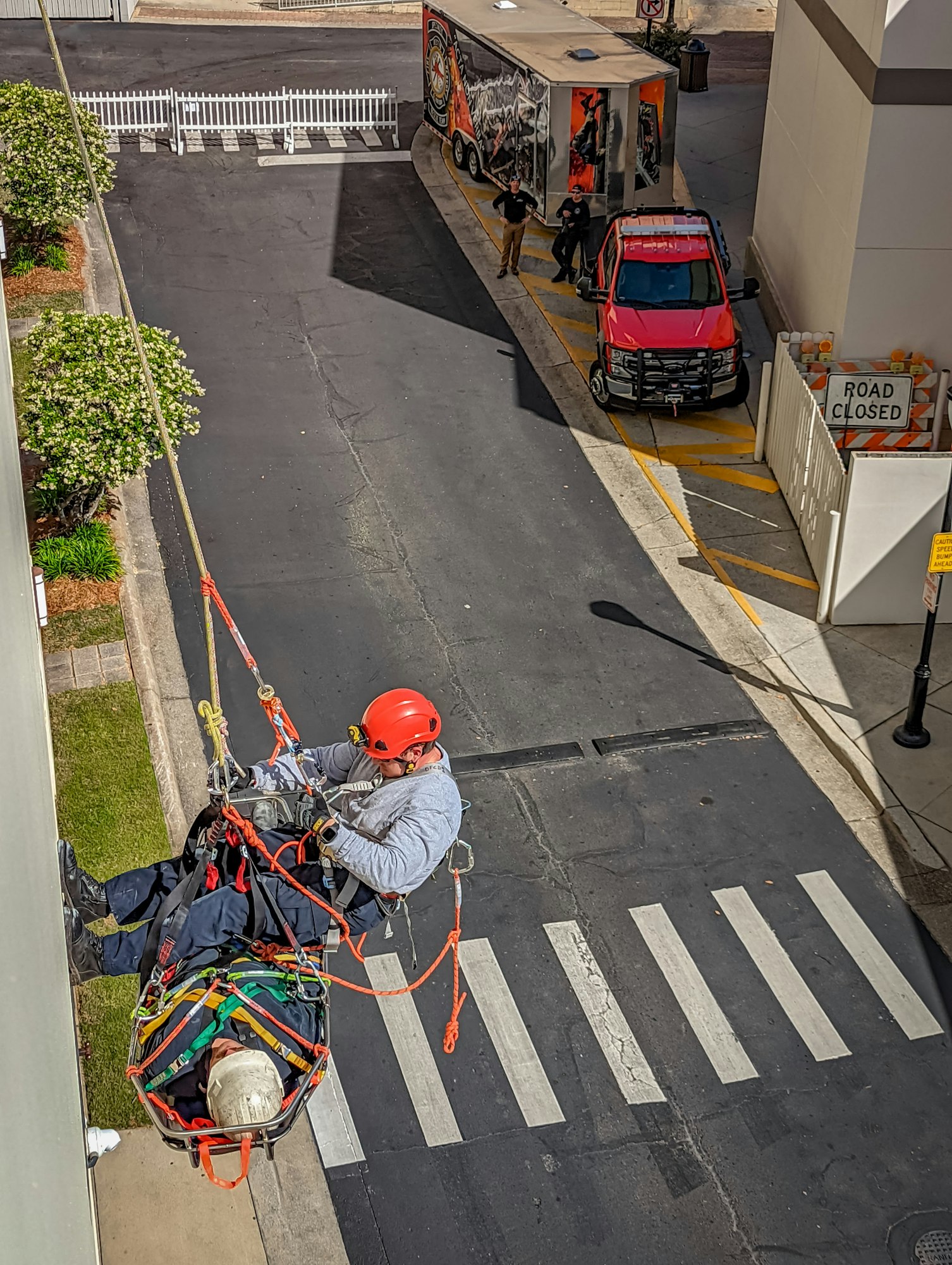 A worker in a harness carries a person on a stretcher down a building with a rope while two people stand by a red vehicle below.