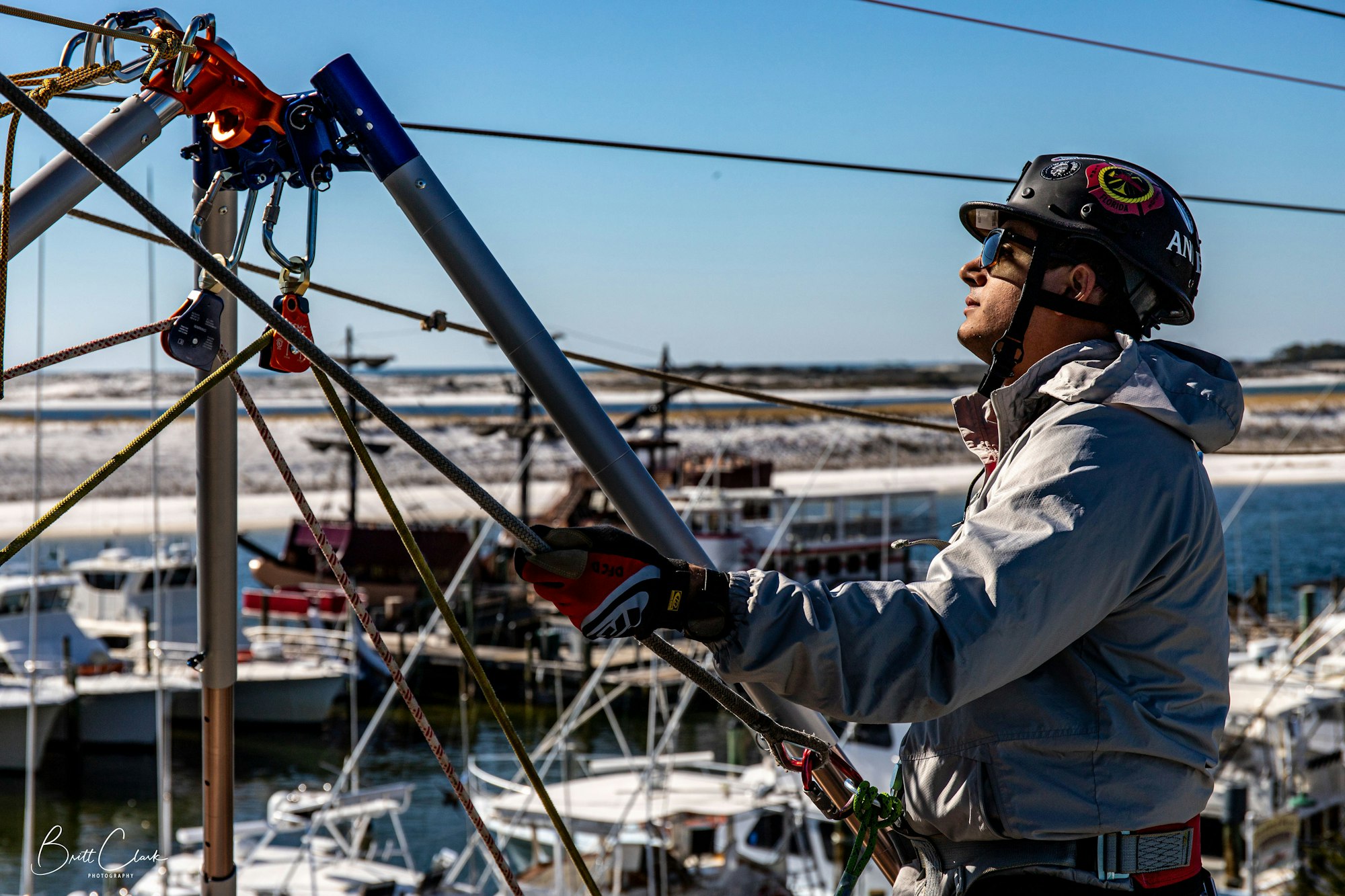 A person wearing a helmet and gloves is adjusting ropes on a structure near a marina.