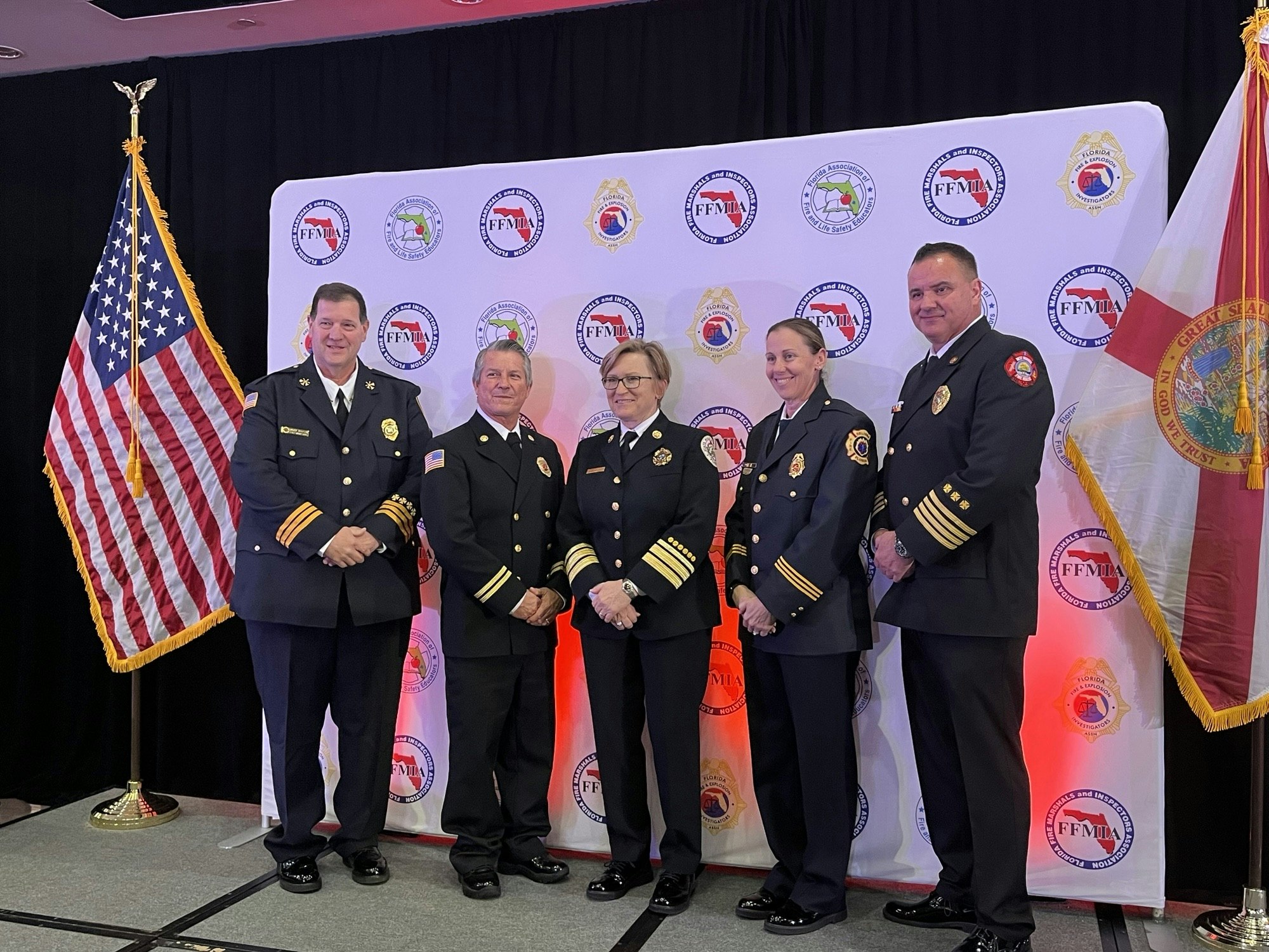 Five people in formal uniforms posing with US and state flags against a backdrop with official logos.