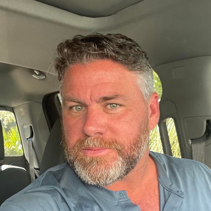 A person with a beard and short hair seated inside a vehicle, wearing a blue shirt.
