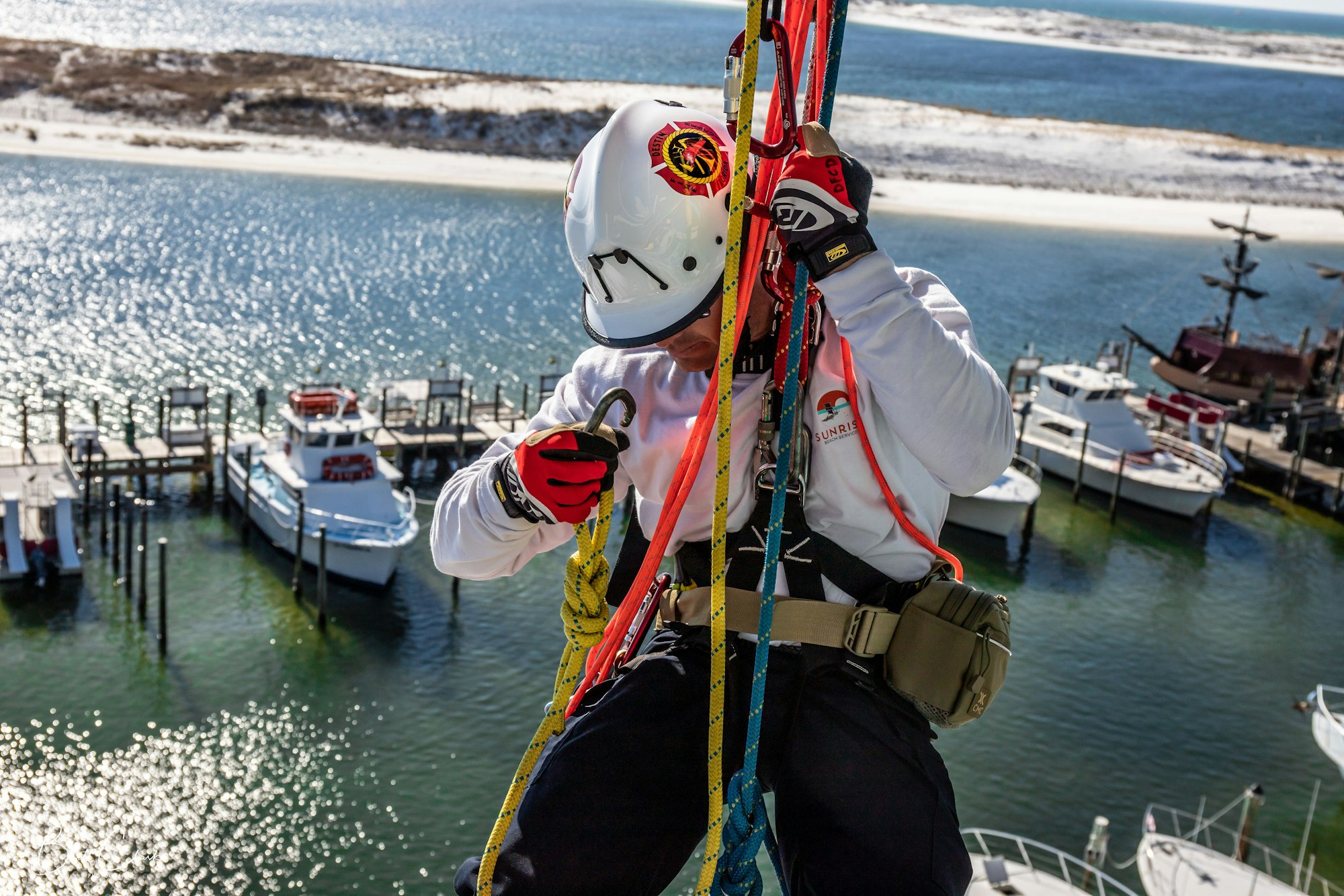 A person in safety gear is performing a rope access task above water with boats in the background.
