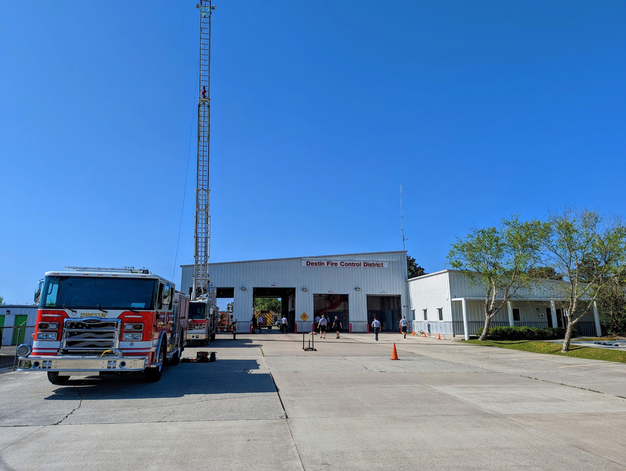 Fire station with a fire truck, extended ladder, and people walking around. Clear blue sky.
