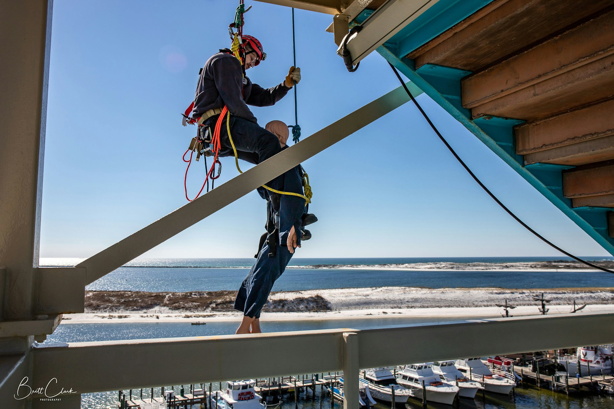 A person wearing safety gear performs a rescue drill, rappelling under a structure near a waterfront with boats docked below.