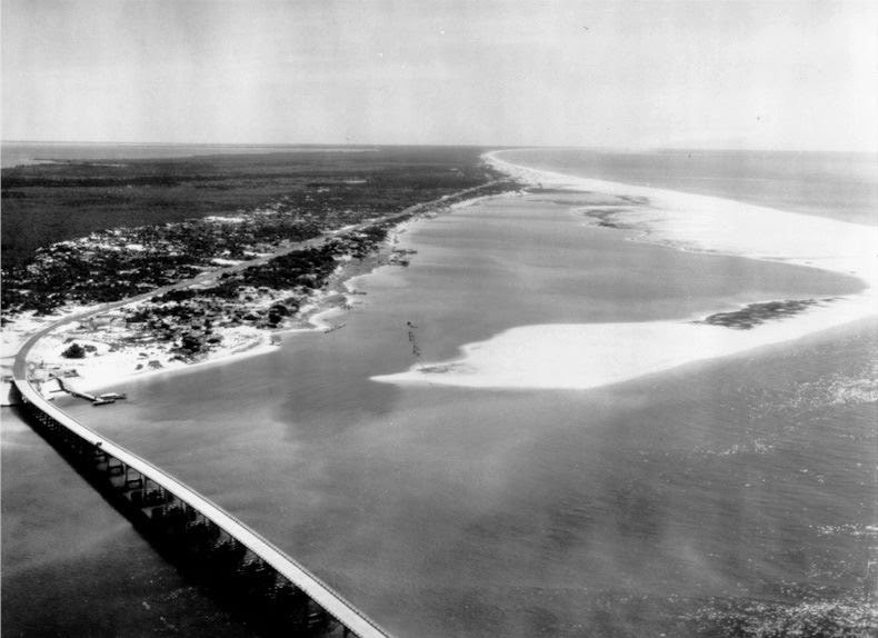 Aerial view of a coastline featuring a bridge, sandy beach, and a vast body of water, captured in black-and-white.