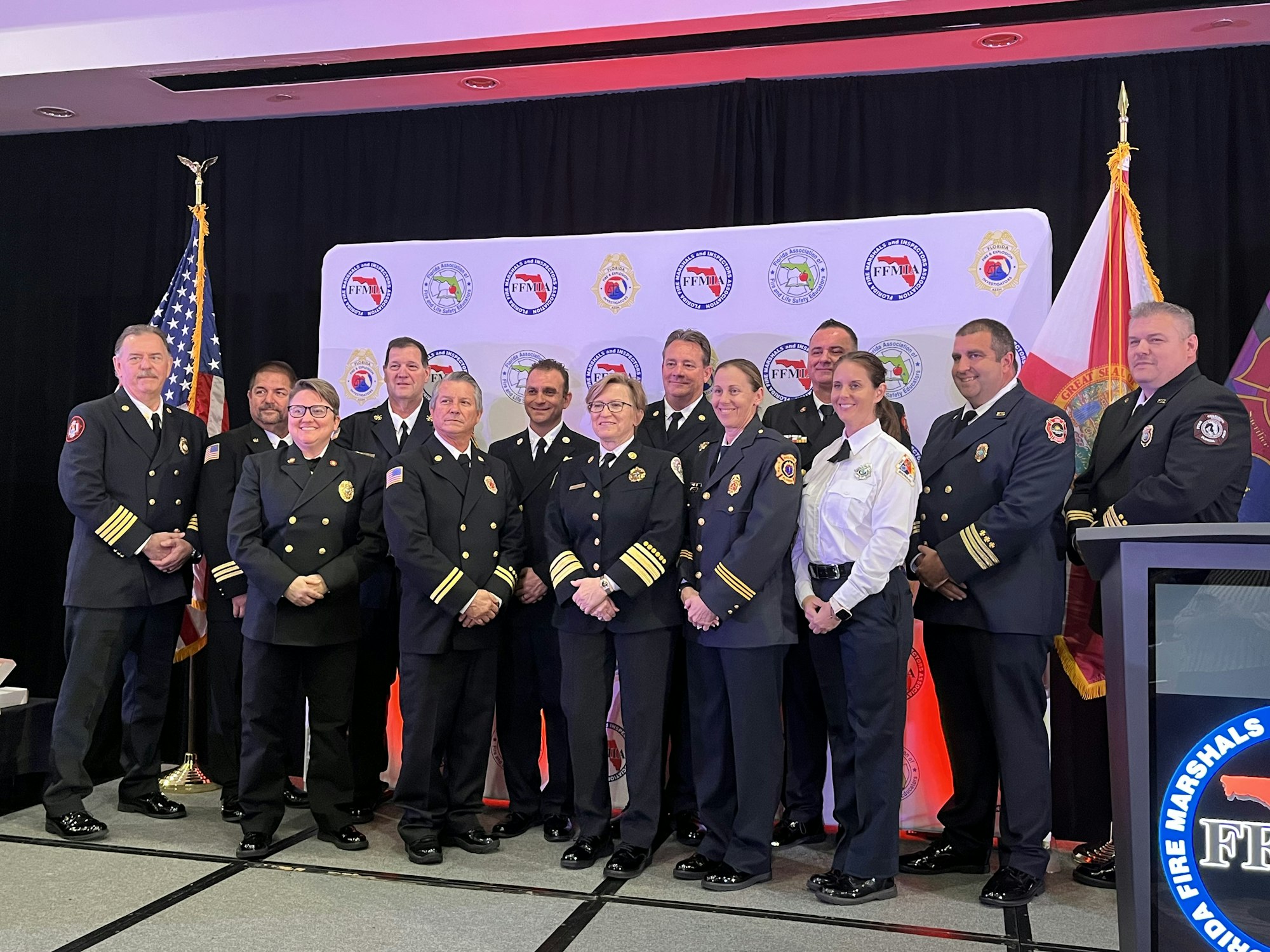 A group of uniformed officials poses for a photo at an event with banners and flags in the background.