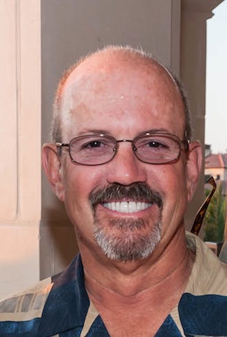 A man with glasses and a beard, smiling, wearing a collared shirt.