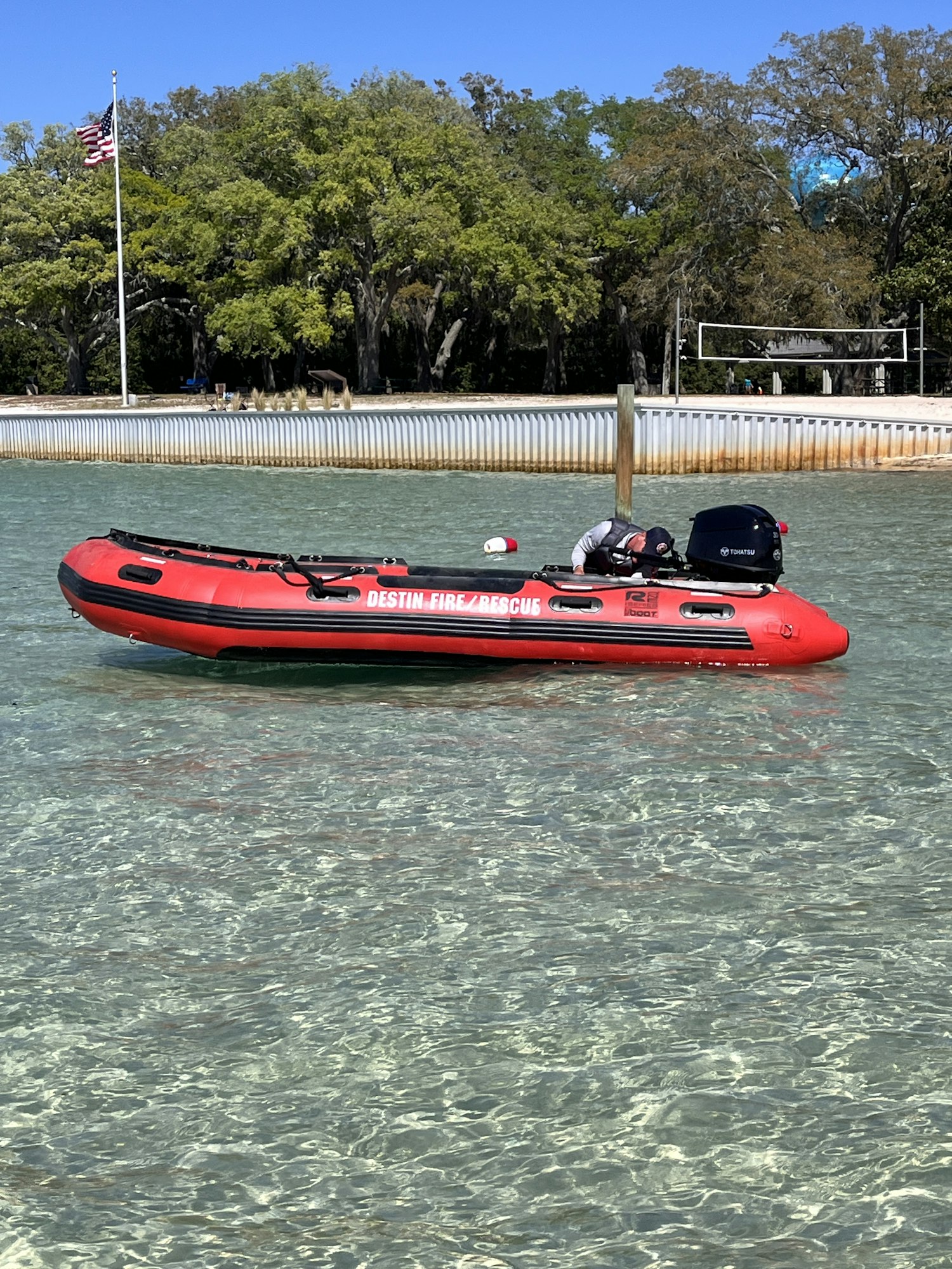 A red inflatable rescue boat on calm water near a dock, with trees and an American flag in the background.
