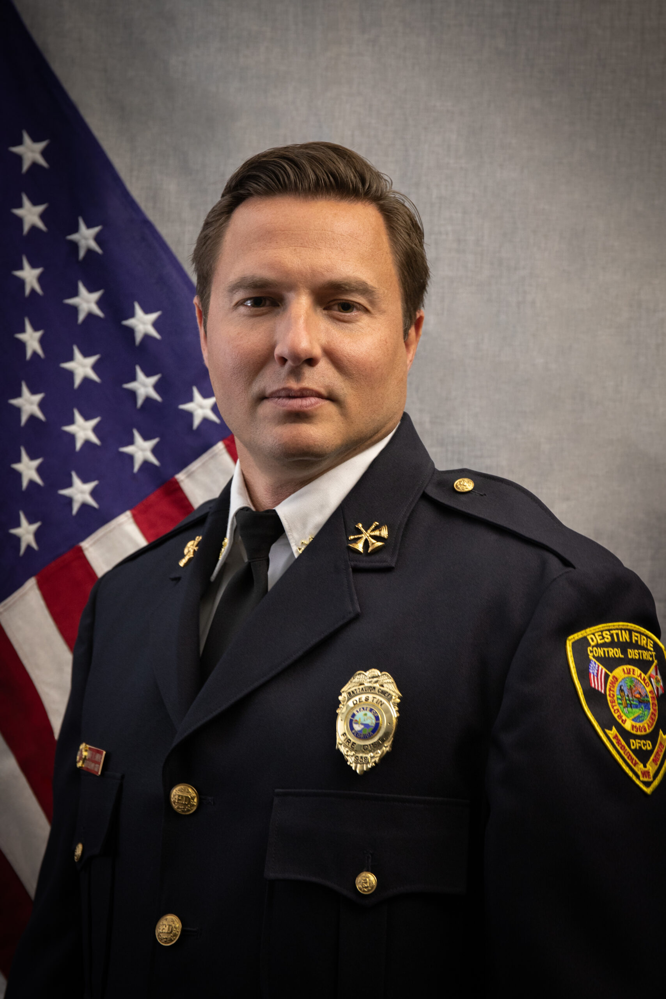 A person in a dark uniform with badges stands before an American flag backdrop.