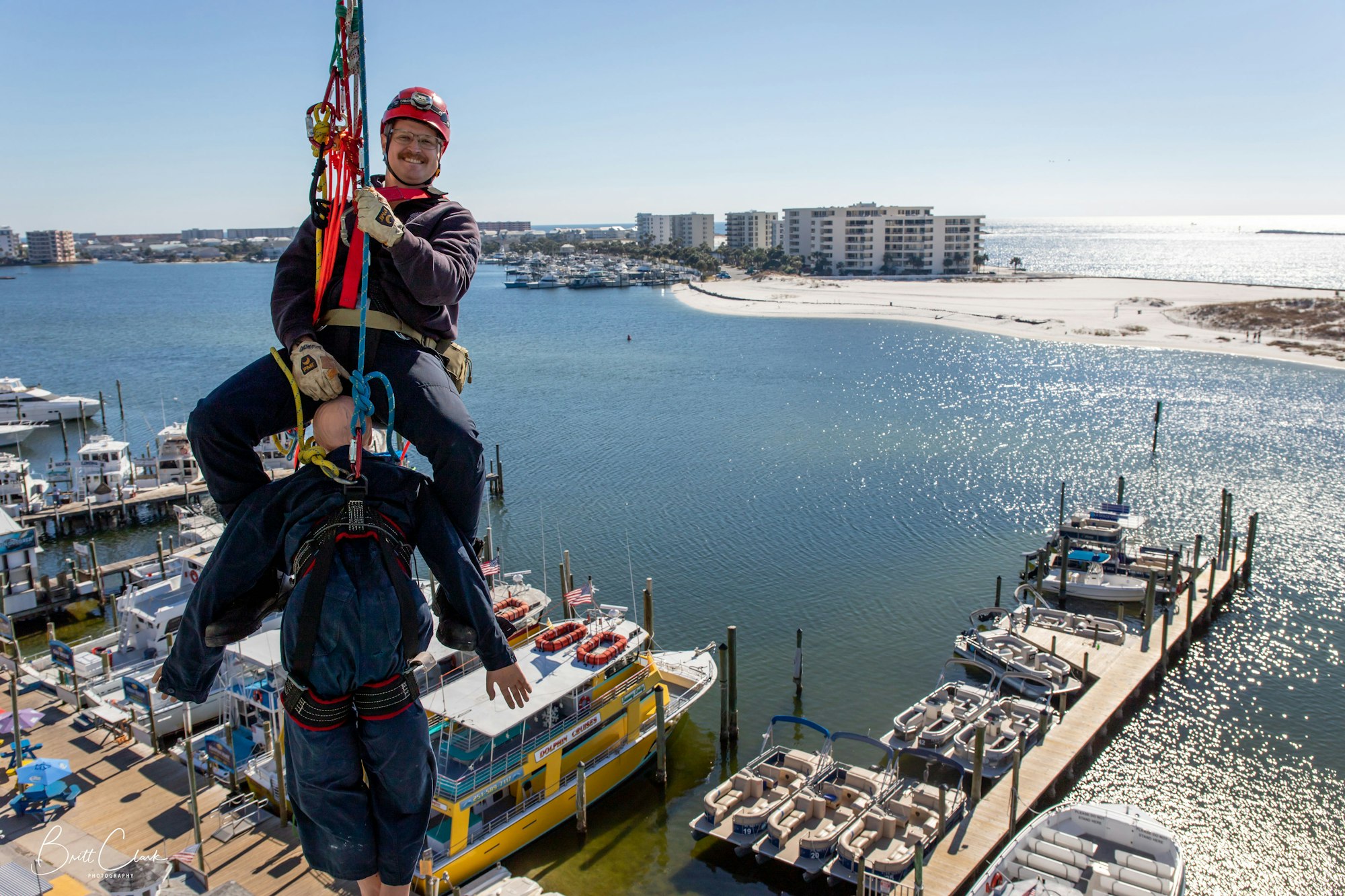 A person in a harness hanging above a marina, practicing rescue techniques with a dummy, against a coastal backdrop.