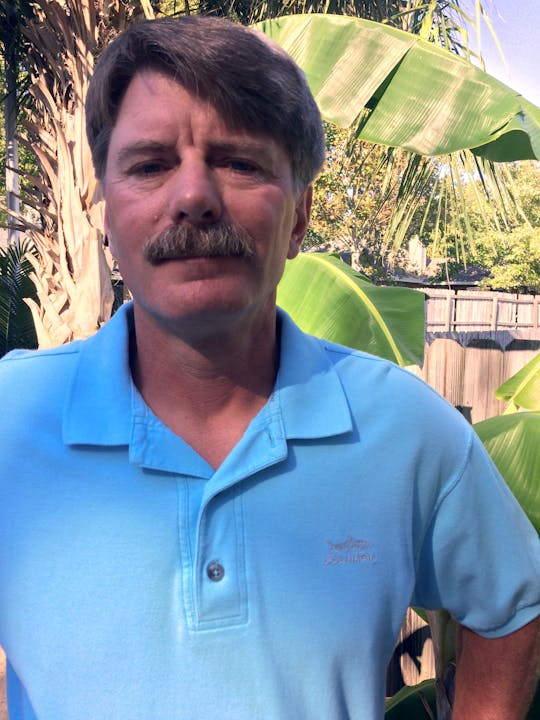 A person in a light blue polo shirt stands outdoors in front of large green leaves.