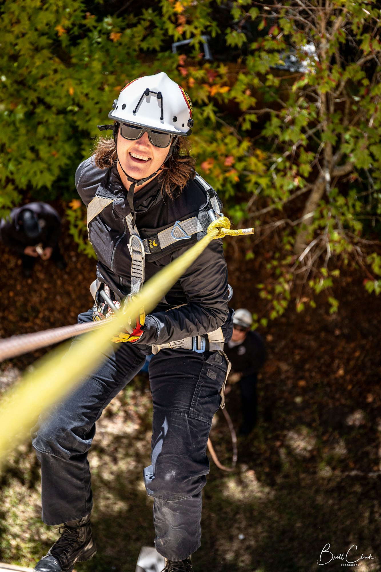 A person rappelling down with safety gear and helmet, surrounded by trees.