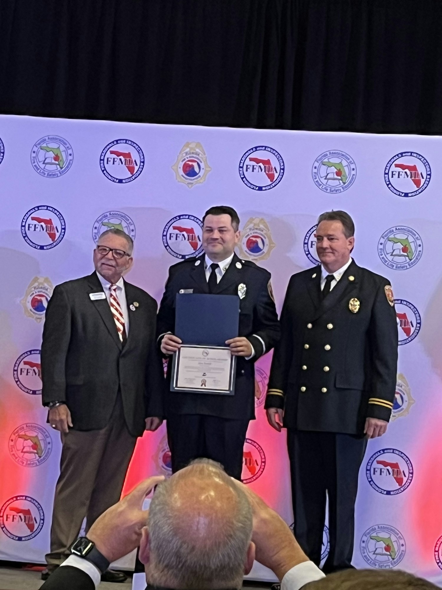 Three men posing at an award ceremony, one holding a certificate, in front of a backdrop with logos.