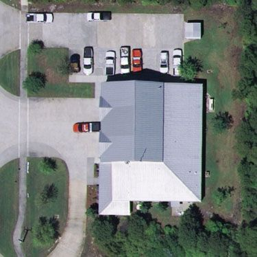 Aerial view of a building with parked cars, surrounded by greenery and a road.