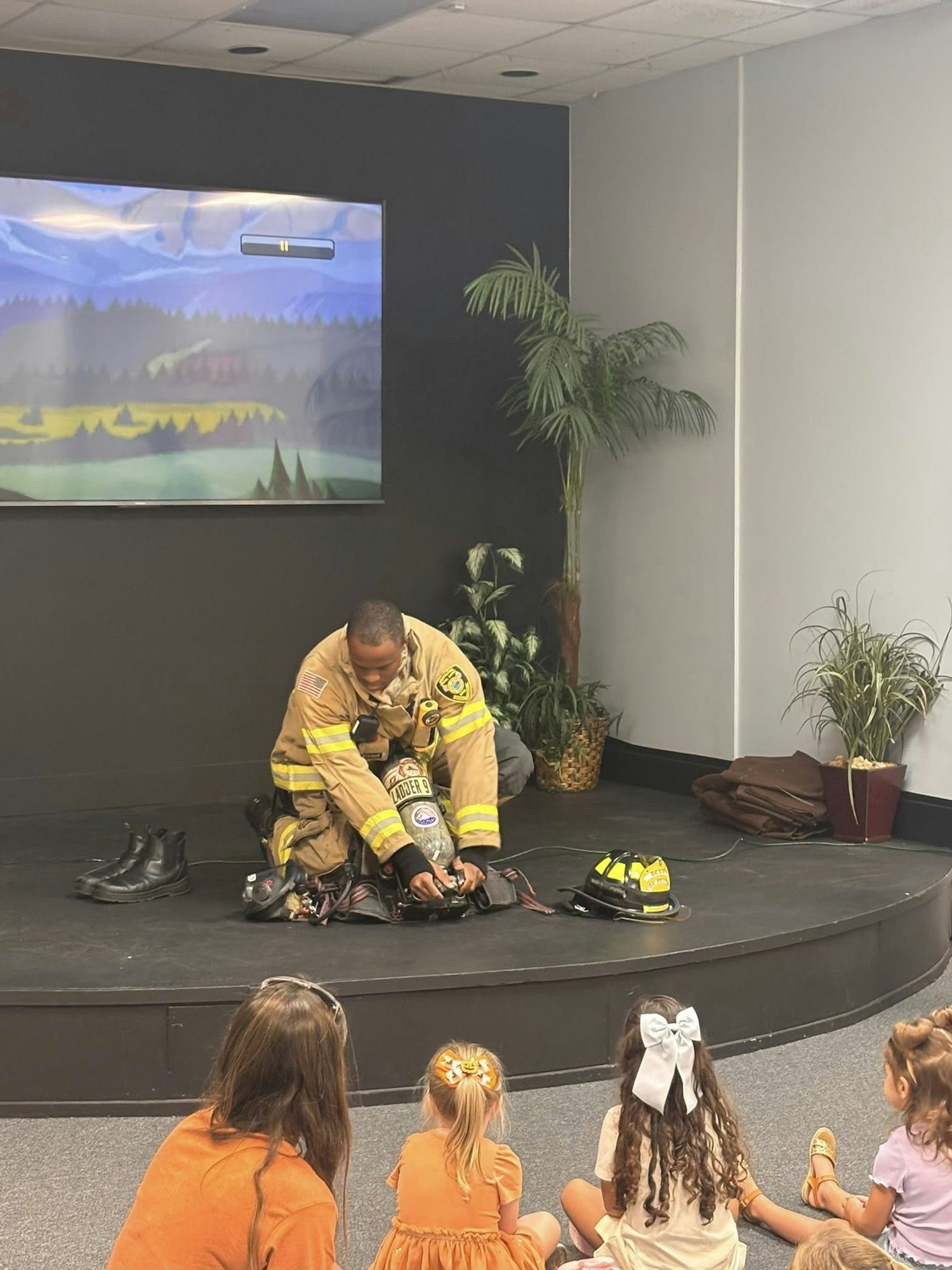 A firefighter demonstrates equipment on stage to a group of children sitting on the floor.