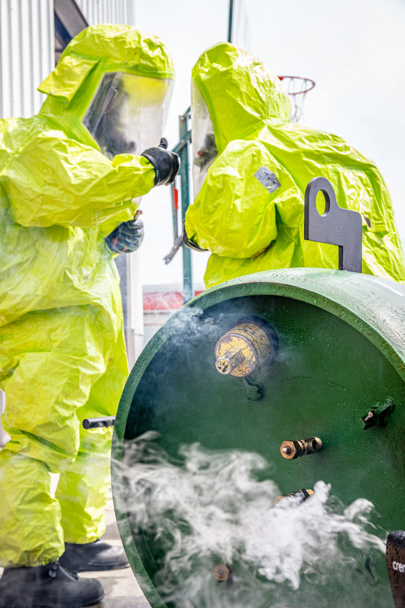 People in hazmat suits inspecting a green cylinder with smoke or gas leaking near it.