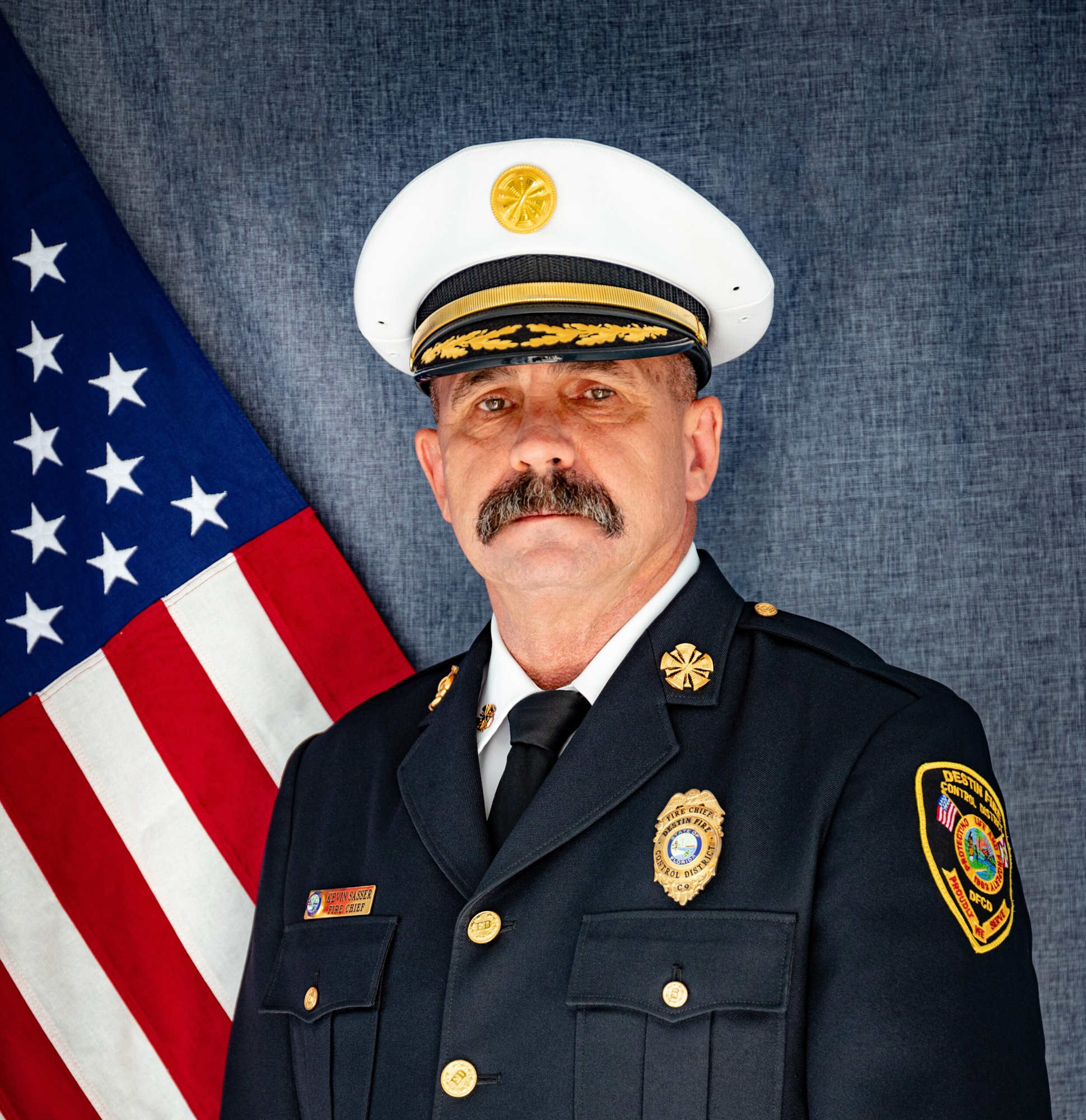 A uniformed fire chief stands in front of an American flag, wearing a white cap with gold accents.