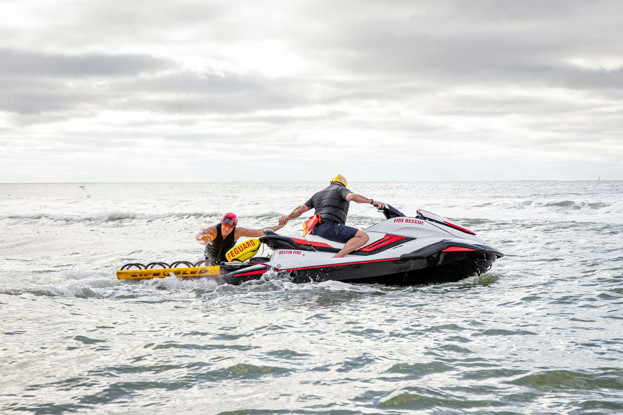 Two people on a rescue jet ski in the ocean, one marked as a lifeguard, another on a "Fire Rescue" vehicle.