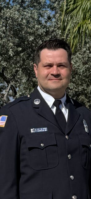 A person in a police uniform with badges stands outdoors in front of trees.