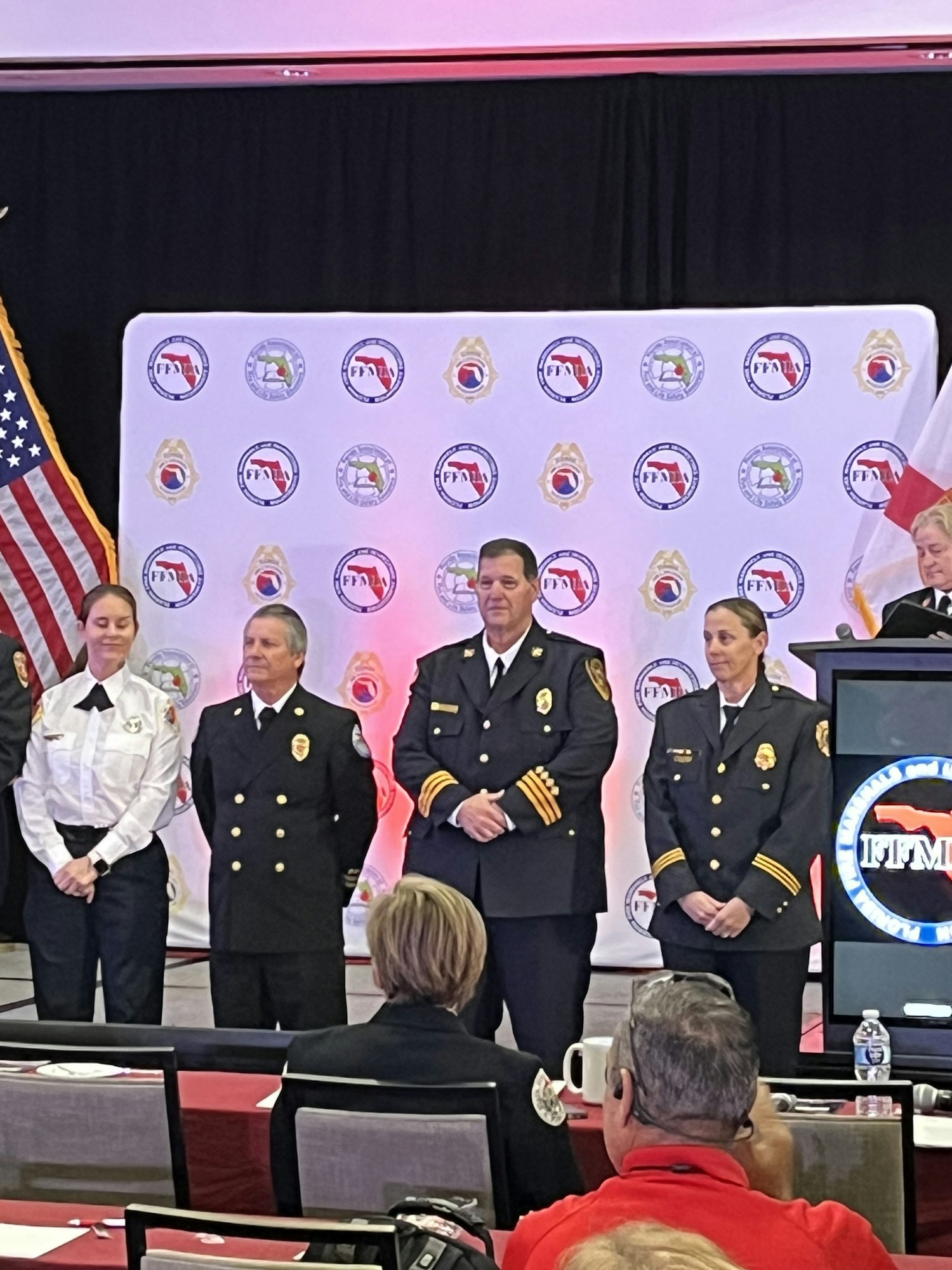 A group of uniformed officials at an event, standing in front of a branded backdrop with logos and an American flag.