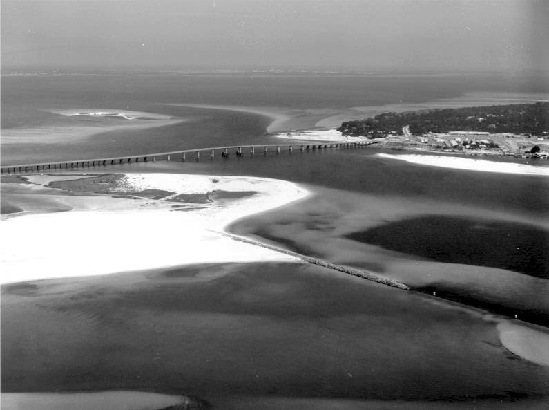 Aerial view of a shoreline with a bridge, sandy areas, and calm waters, captured in black and white.