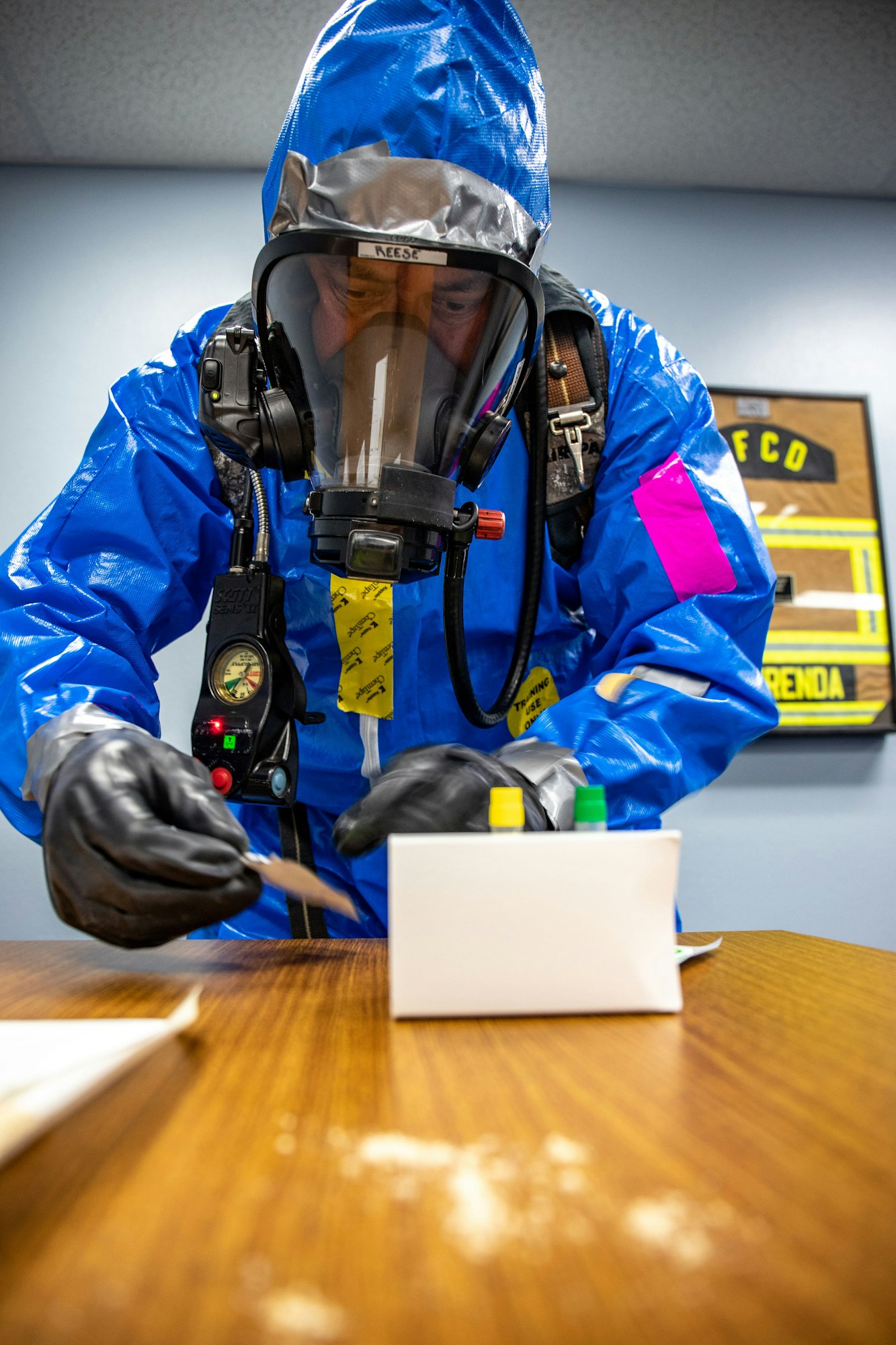 Person in hazmat suit, gloves, and respirator inspecting powder on a table.