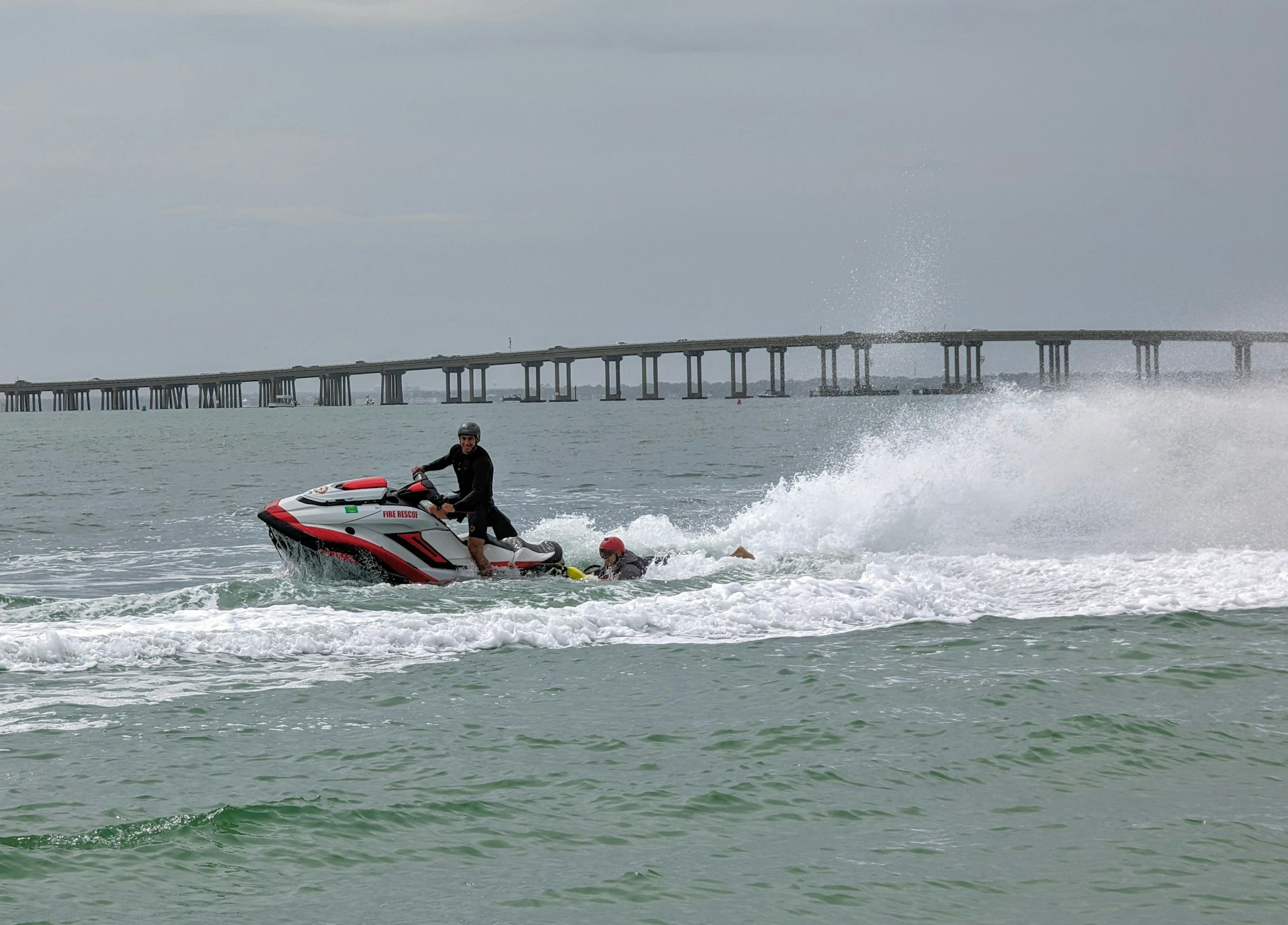 A person on a jet ski labeled "Fire Rescue" in the ocean, with a bridge in the background.