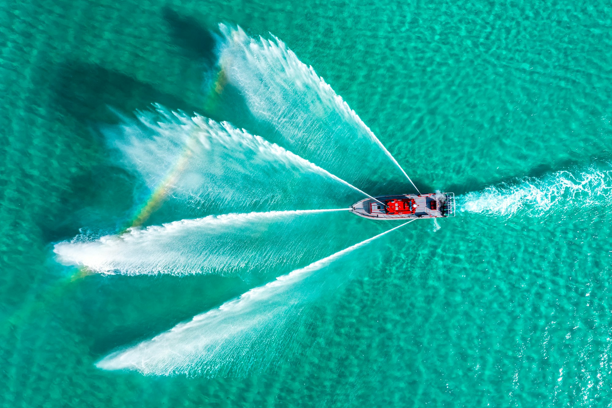 Aerial view of a boat on turquoise water, creating symmetric water sprays, with a faint rainbow visible.