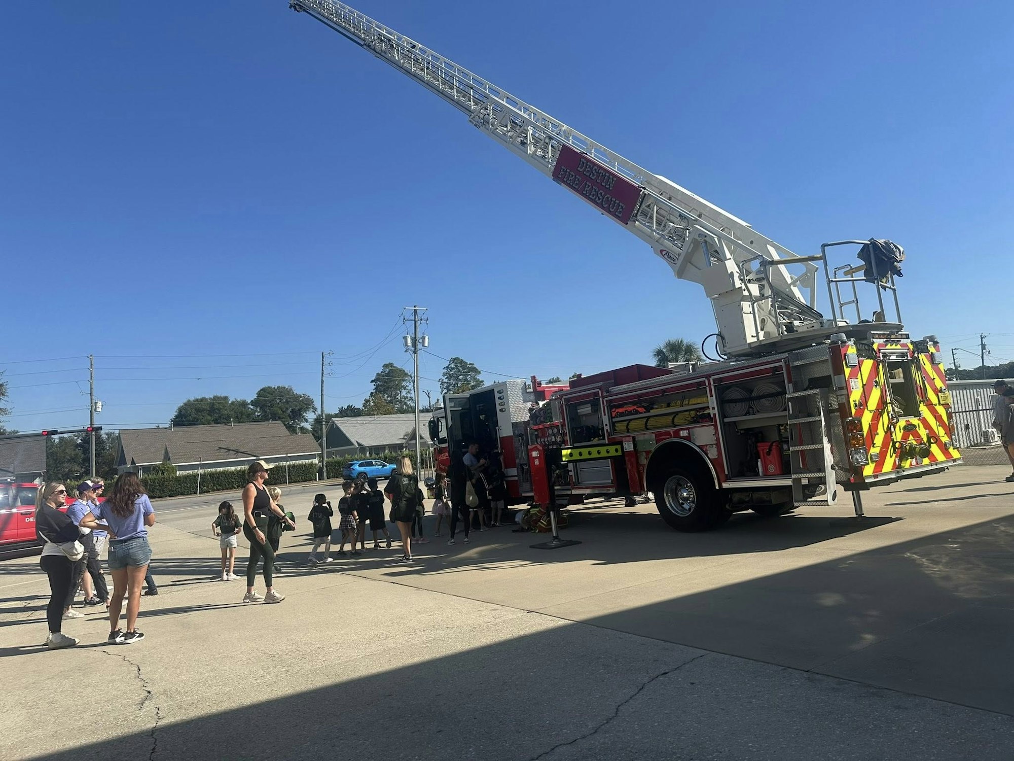 A fire truck with an extended ladder is parked, surrounded by people on a sunny day.