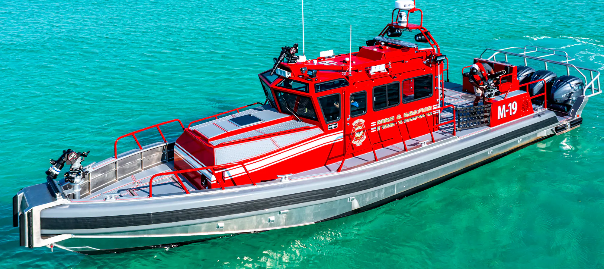 A red fire and rescue boat labeled "M-19" in clear blue water with a bridge and shoreline in the background.