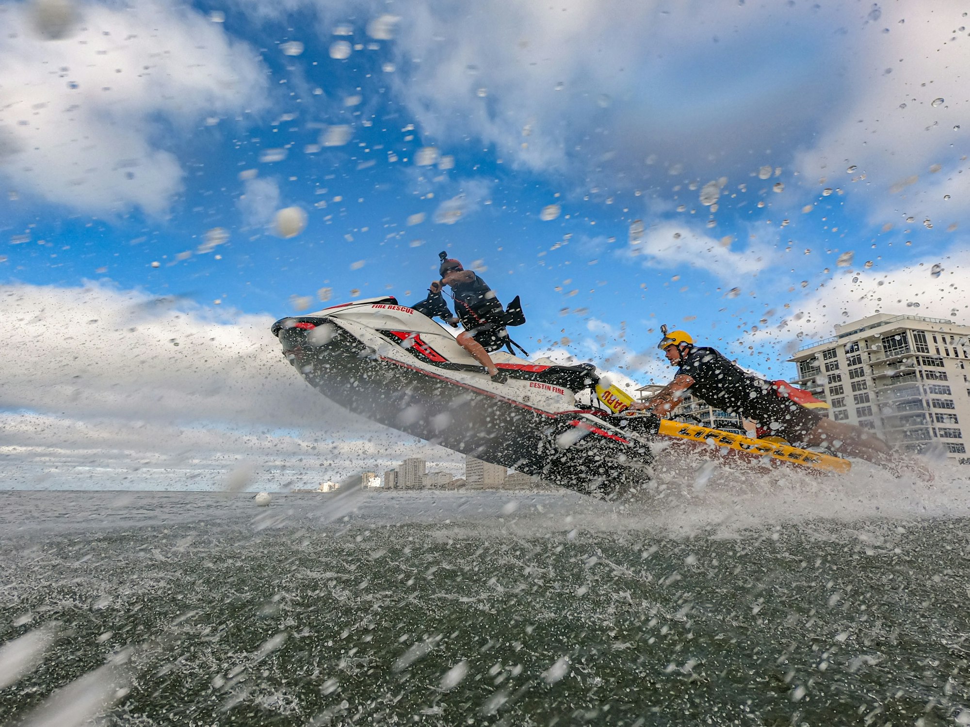 Two people on a jet ski jumping over waves, with splashes of water around them against a backdrop of buildings and blue sky.