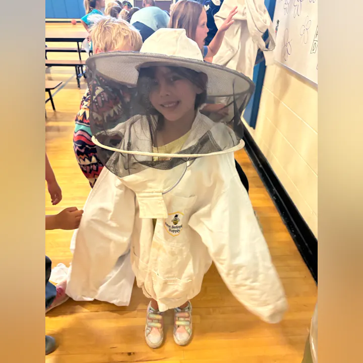 A child in a bee suit and hat smiles, surrounded by peers in a school setting.