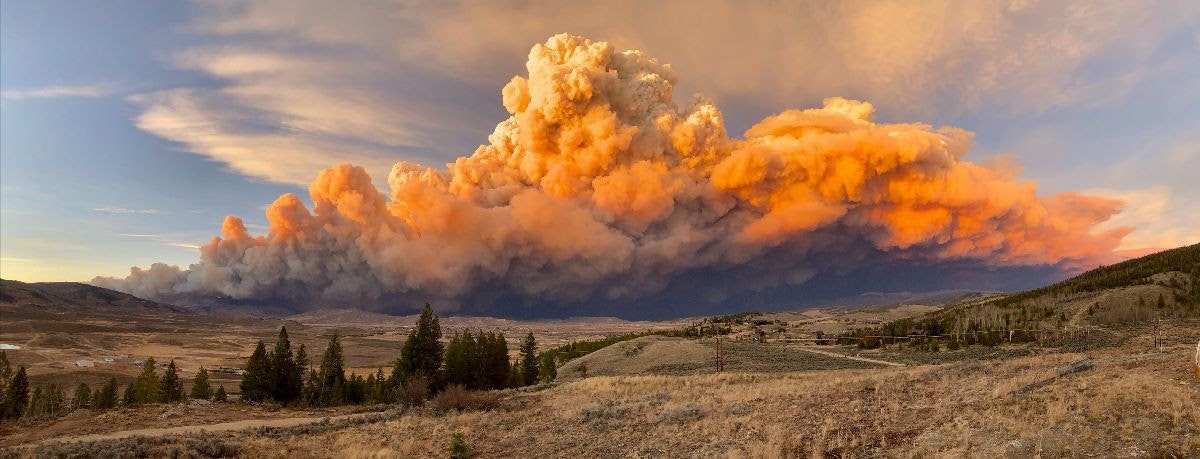 A vast cloud of smoke from a wildfire looms over a rural landscape during sunset, with orange hues illuminating the sky.