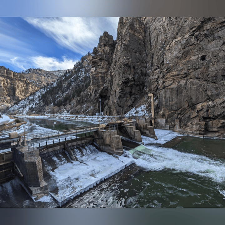The image shows a dam on a river surrounded by rocky cliffs and some snow, with flowing water and a clear blue sky.
