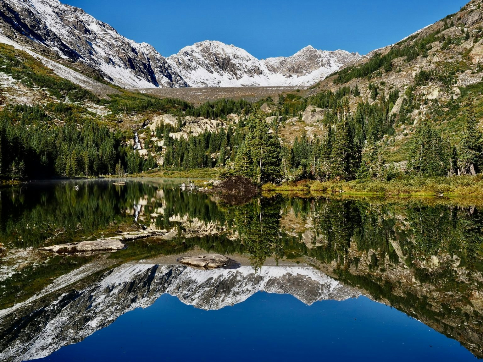 A serene landscape featuring majestic mountains, lush trees, and a calm lake reflecting the scenery beneath a clear blue sky.