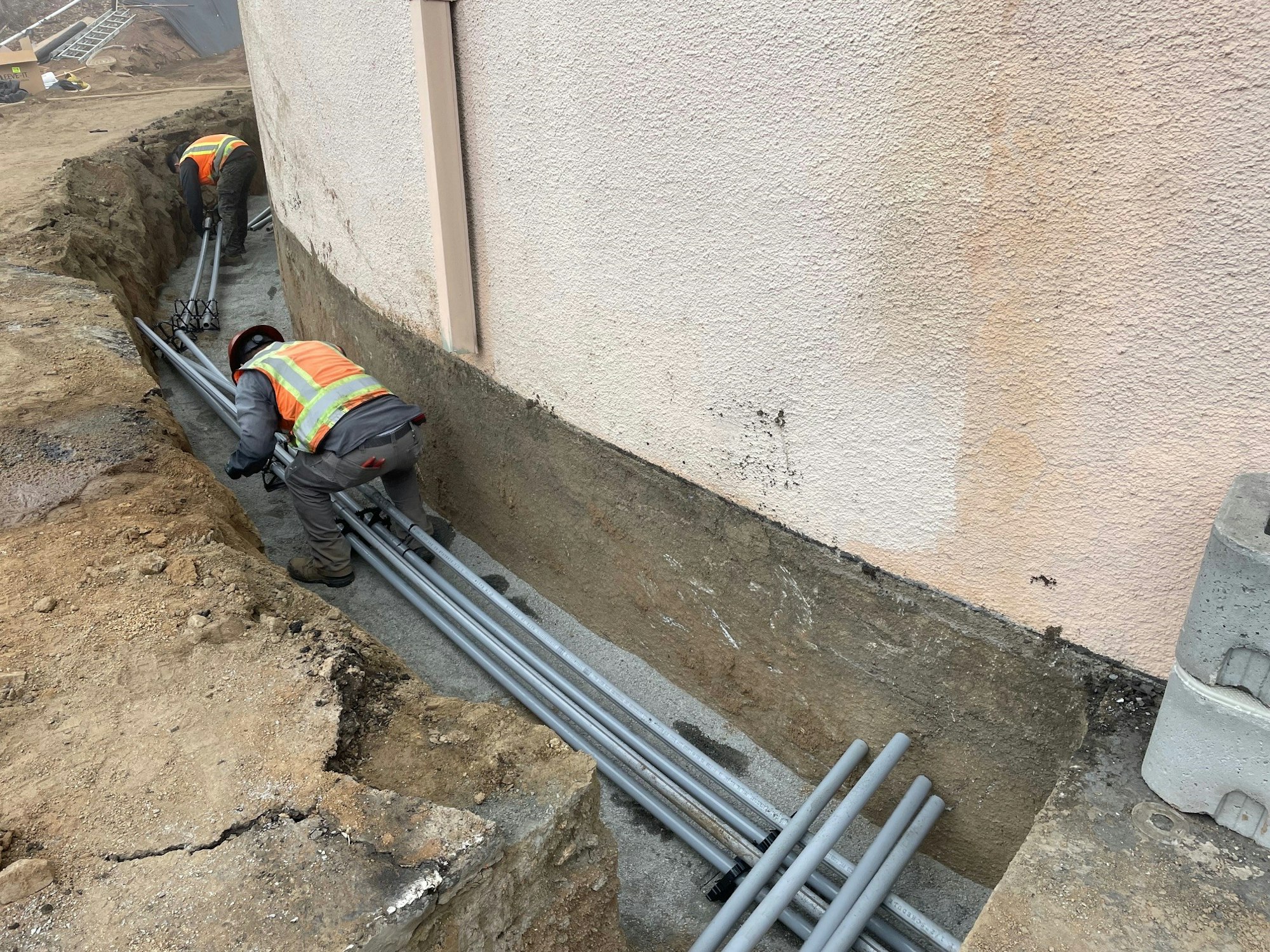 Construction workers installing metal pipes in a trench beside a building wall.