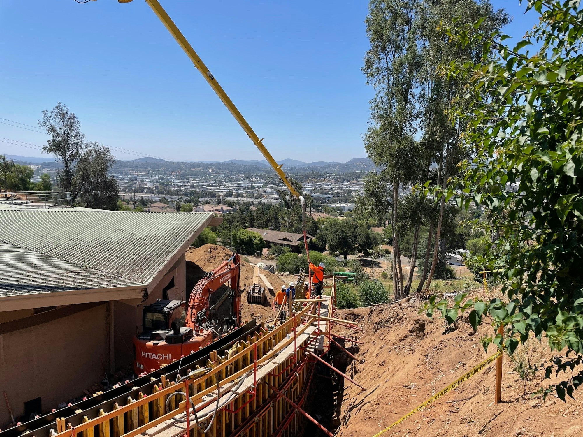 Construction site with a Hitachi excavator, retaining wall setup, overlooking a cityscape and hills.