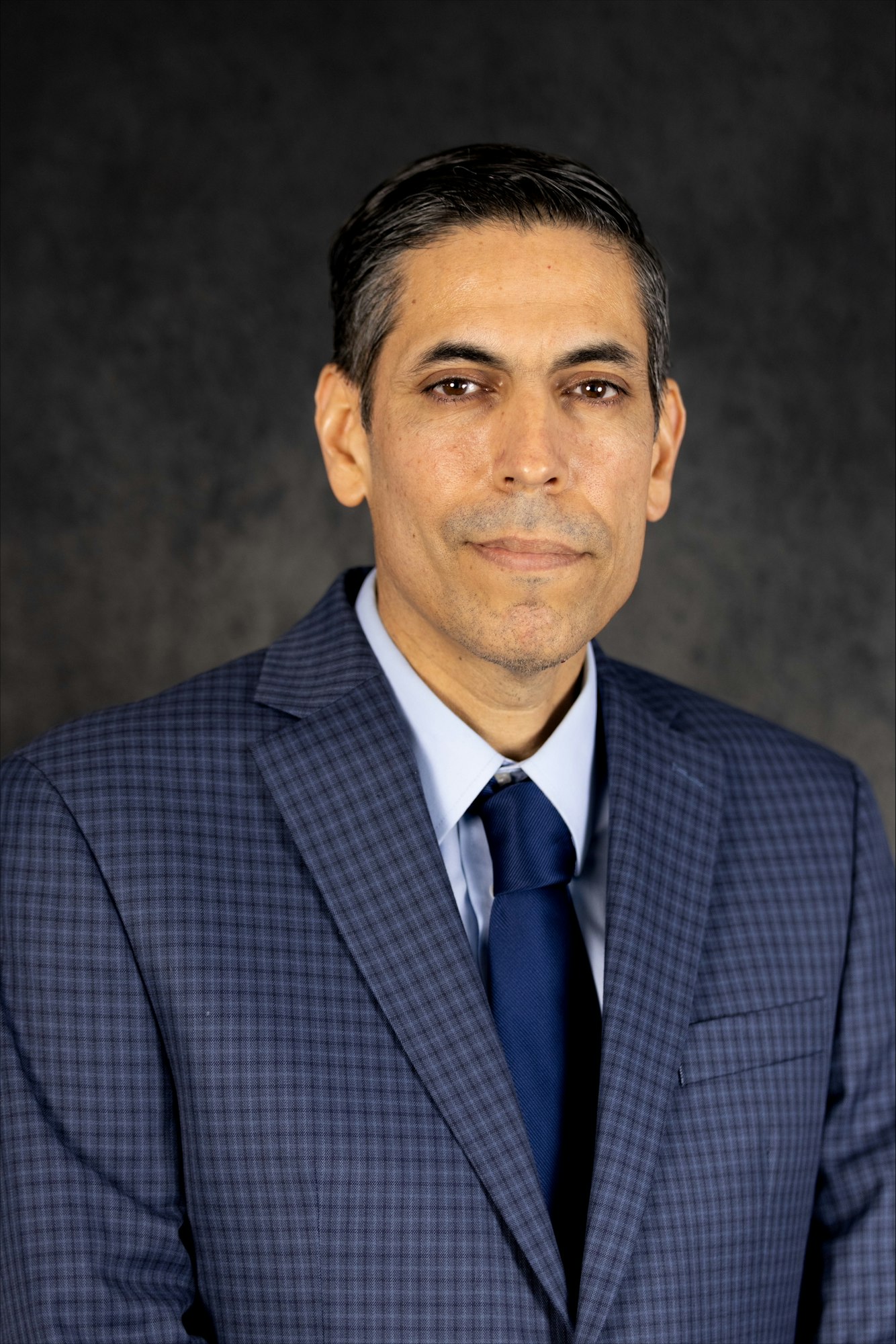 A person in a blue checkered suit and tie against a dark background.