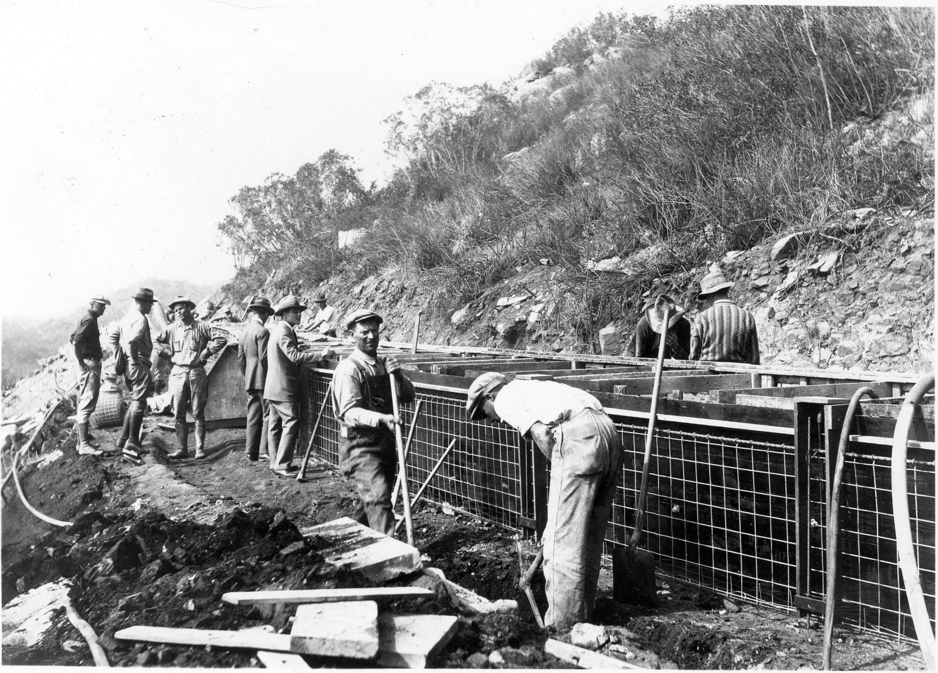 Workers constructing a concrete form along a hillside, using shovels, with some overseeing the progress.