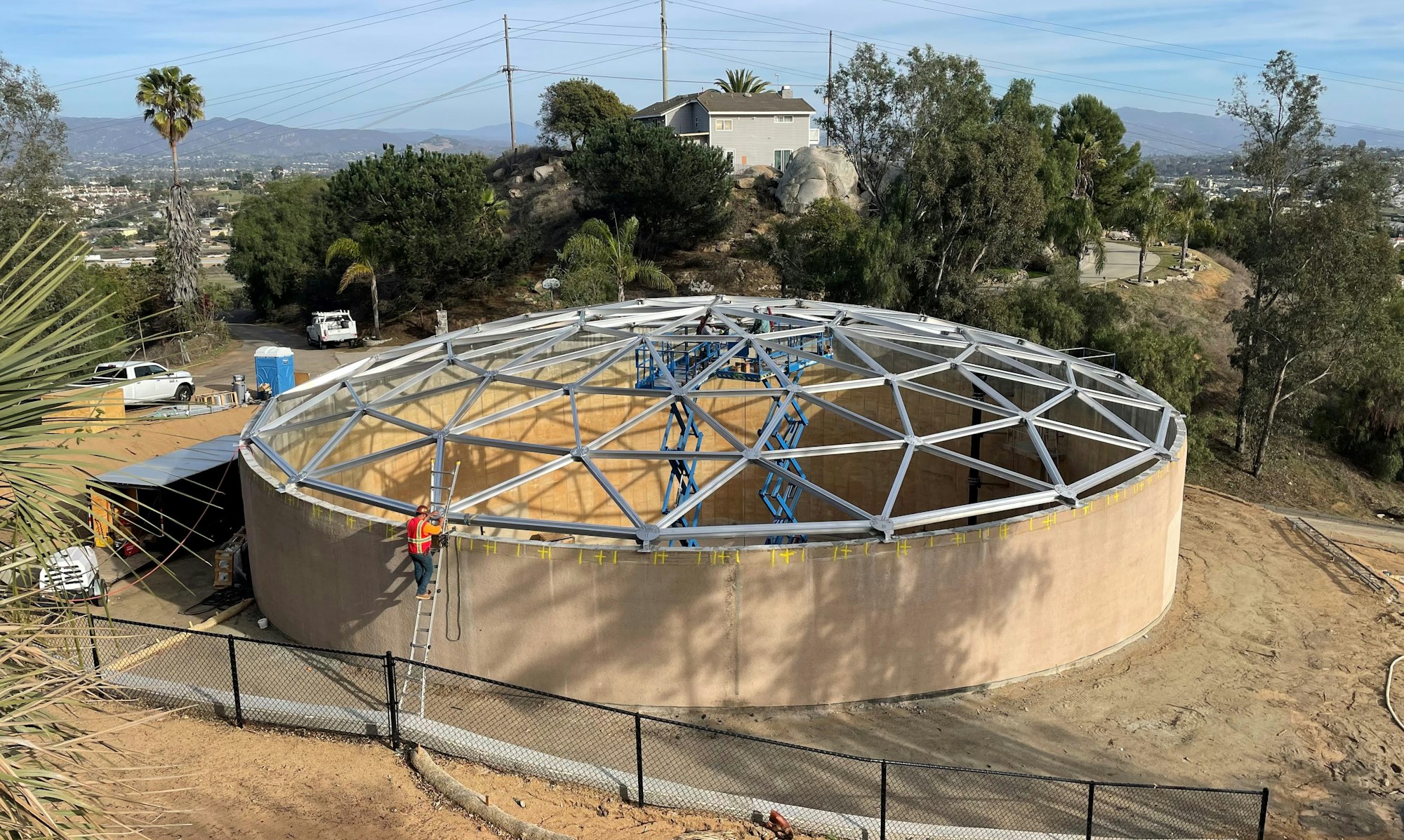 A construction site with a circular structure, a geodesic dome frame, workers, and vehicles in a hilly landscape.