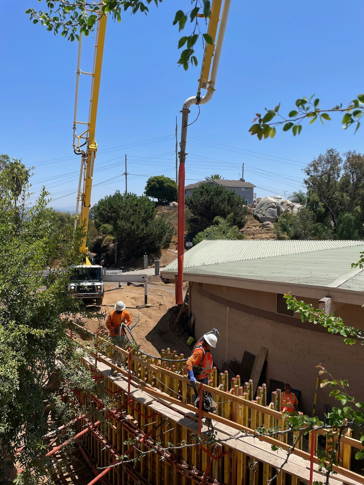 Construction workers in orange gear pour concrete into a form with a long boom pump, outdoors under a clear blue sky.