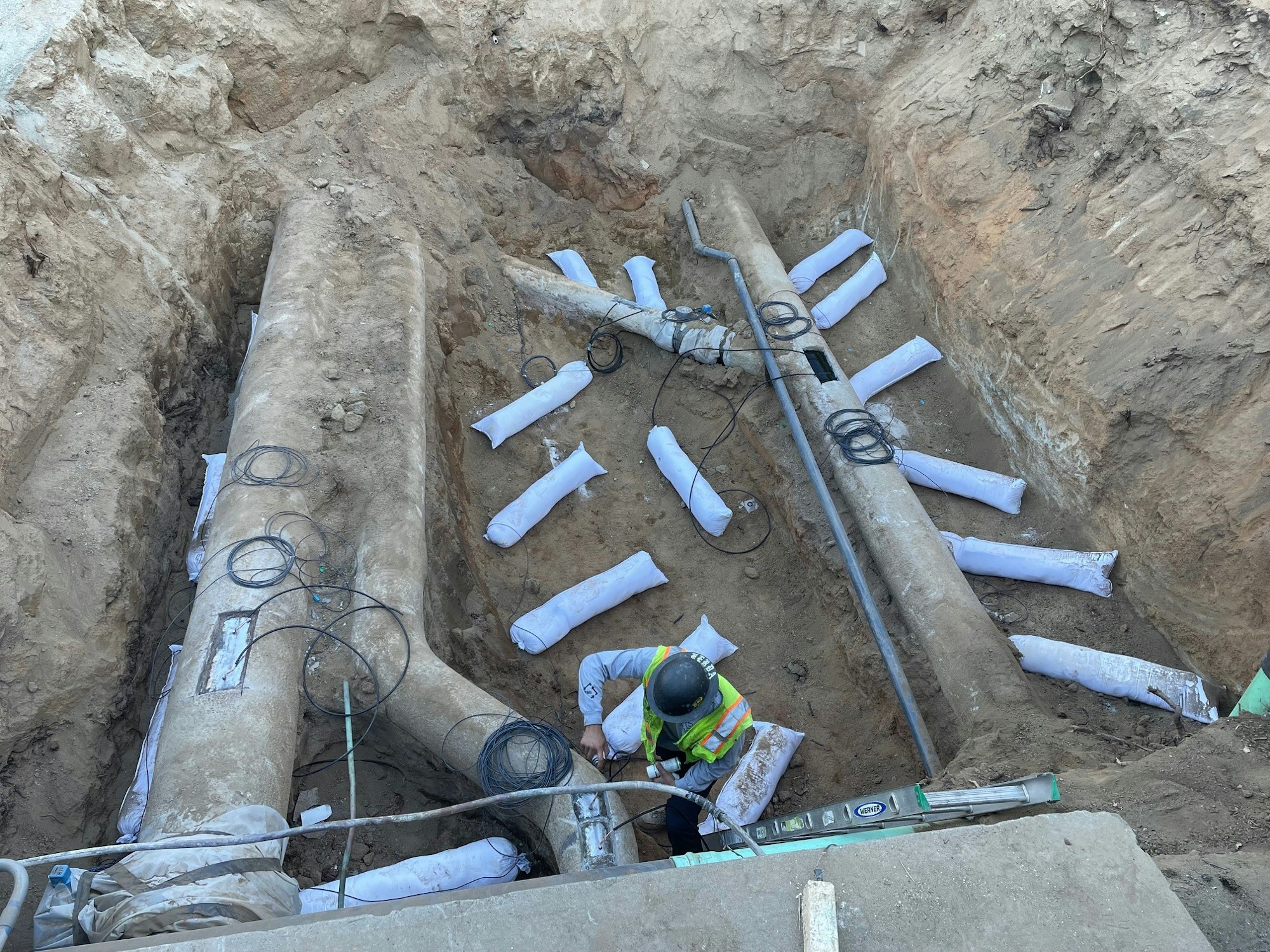 Construction worker inspecting large pipes in a trench with sandbags and cables around.